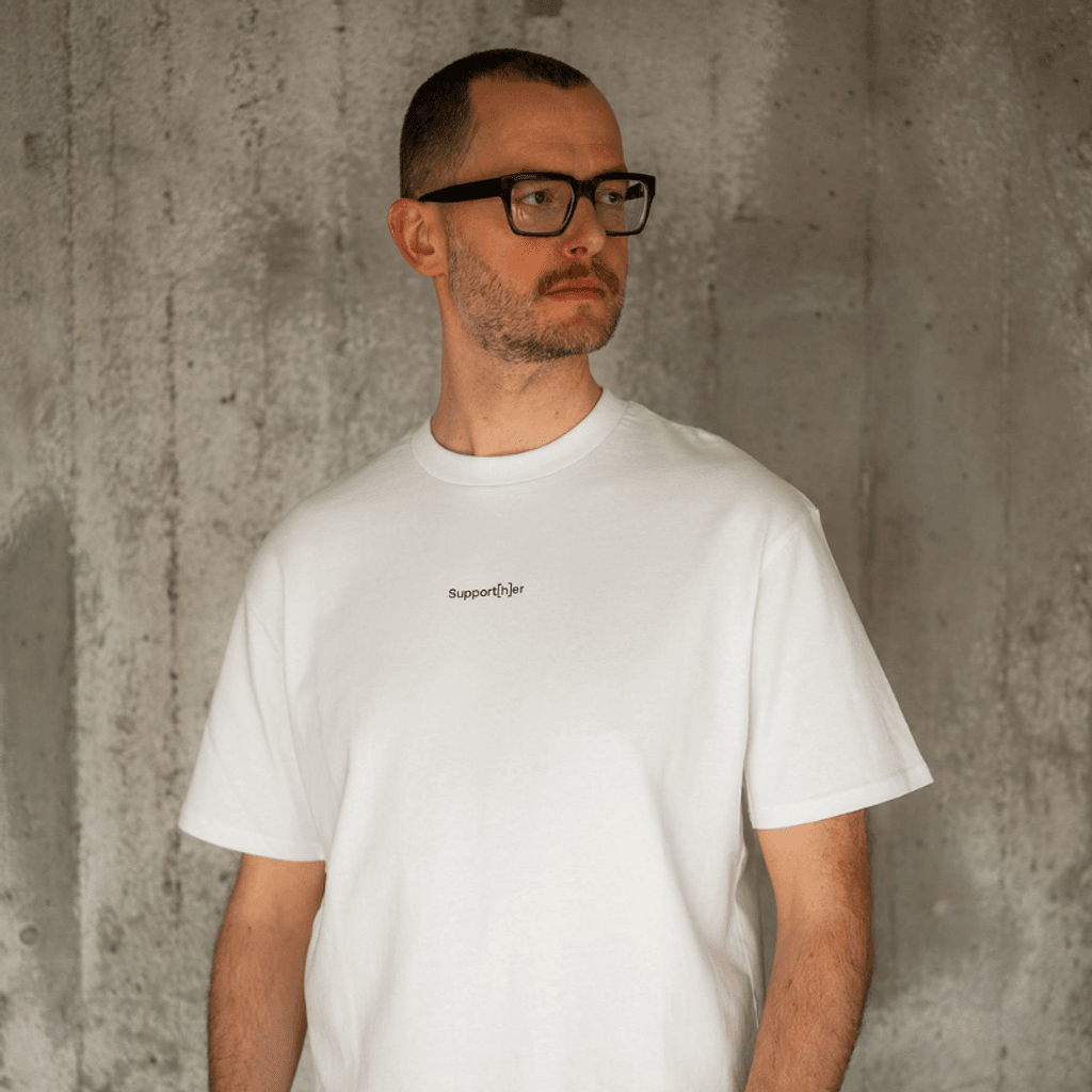 Person trägt weißes T-Shirt mit einem mit einem kleinen Logo Support[h]er auf der Brust