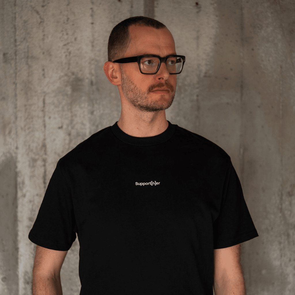 Person trägt schwarzes T-Shirt mit einem mit einem kleinen Logo support[h]er auf der Brust