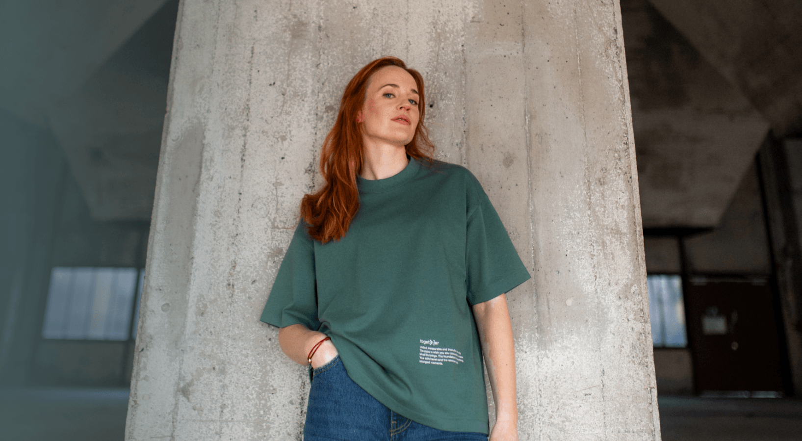 Frau mit roten Haaren trägt grünes T-Shirt mit kleinem Schriftzug unten rechts und steht vor einer Betonwand