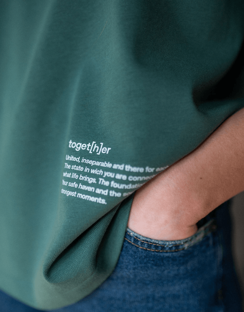 Detailaufnahme eines grünen T-Shirts mit kleinem „toget[h]er“-Schriftzug und Text am unteren Saum, getragen von einer Person mit Hand in der Hosentasche