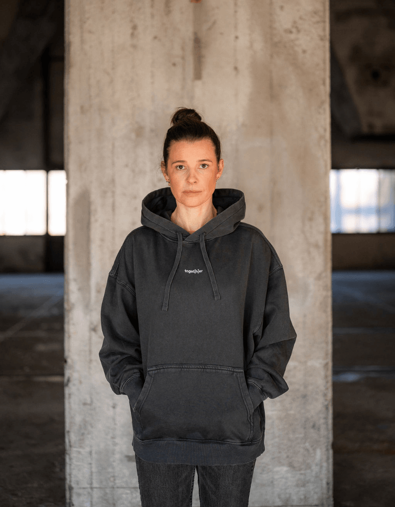 Person trägt schwarzen Hoodie mit kleinem Schriftzug toget[h]er auf der Brust vor Betonwand