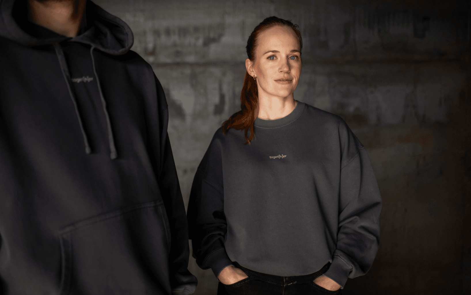 Person trägt dunkelgraues Sweatshirt mit kleinem Logo toget[h]er auf der Brust vor neutralem Hintergrund