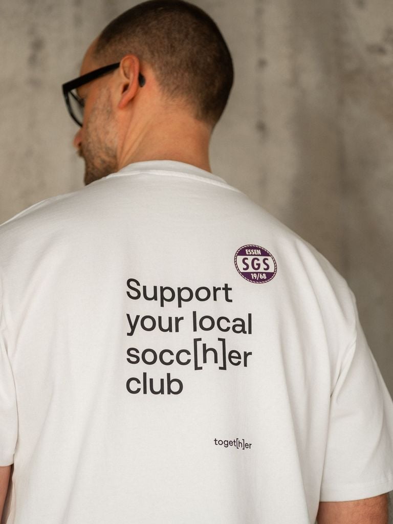 Model trägt weißes unisex T-Shirt mit kleinem Schriftzug toget[h]er auf der Brust. Das T-Shirt ist aus der Kollektion socc[h]er. 