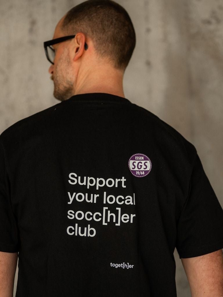 Person trägt schwarzes T-Shirt mit Schriftzug „Support[h]er“ auf der Brust