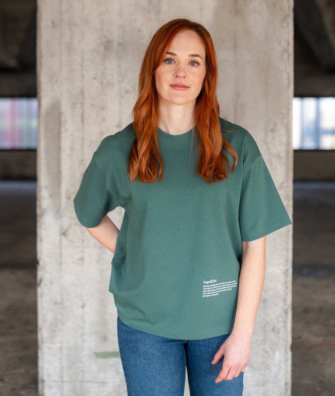 Frau mit roten Haaren trägt ein grünes T-Shirt mit kleinem Schriftzug unten rechts, steht vor einer Betonwand und schaut in die Kamera