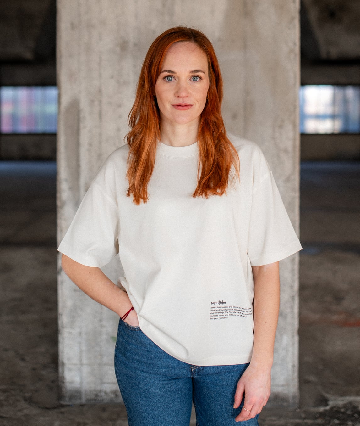 Frau mit roten Haaren trägt ein weißes T-Shirt mit kleinem Schriftzug unten rechts, steht vor einer Betonwand und schaut in die Kamera