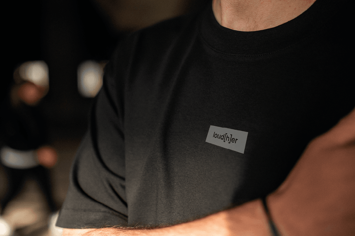 Detailaufnahme einer Person, die schwarzes T-Shirt mit Schriftzug „loud[h]er“ auf der Brust trägt