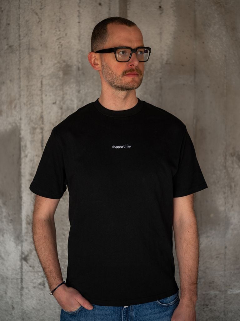 Person trägt schwarzes T-Shirt mit Schriftzug „Support[h]er“ auf der Brust
