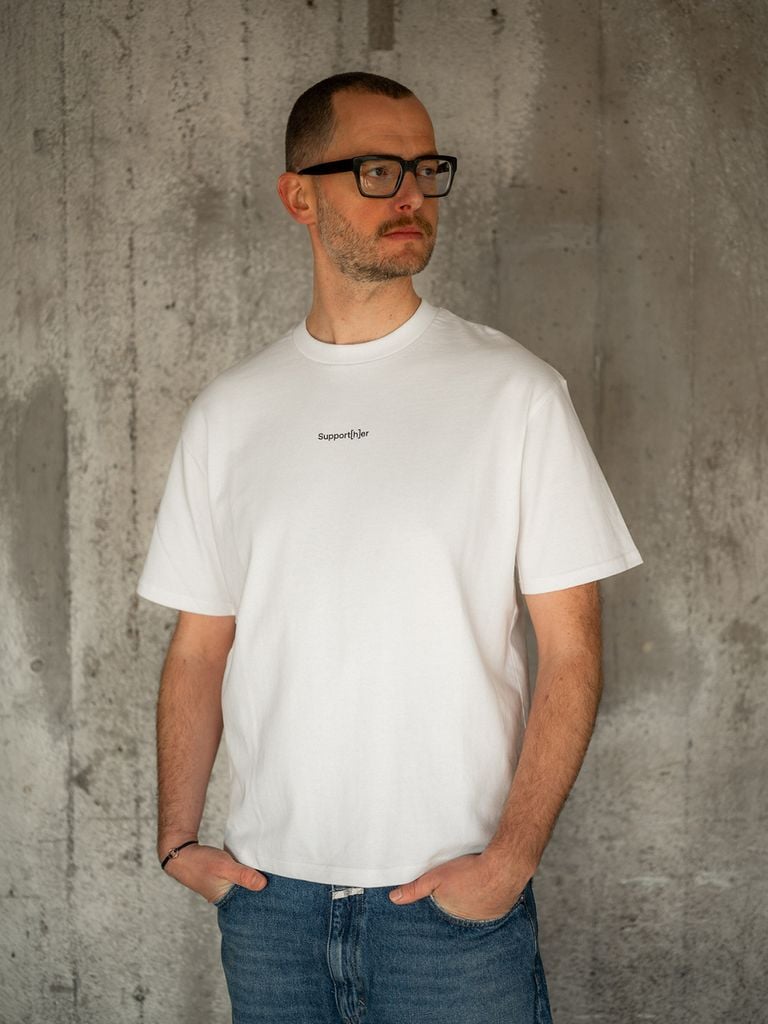 Model trägt weißes unisex T-Shirt mit kleinem Schriftzug toget[h]er auf der Brust. Das T-Shirt ist aus der Kollektion socc[h]er. 