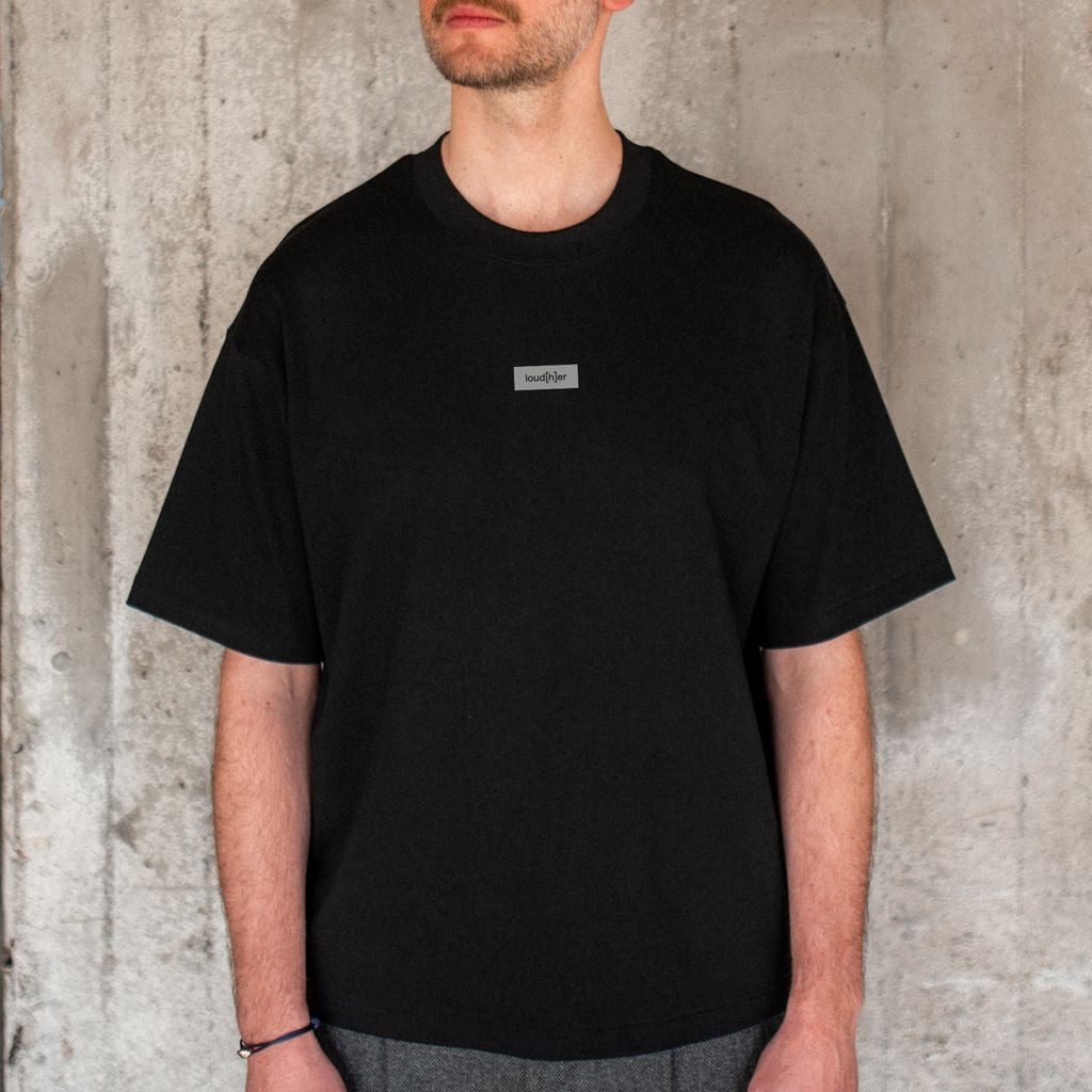 Model trägt schwarzes unisex T-Shirt mit Schriftzug loud[h]er auf der Brust. Das T-Shirt ist aus der Kollektion loud[h]er. 