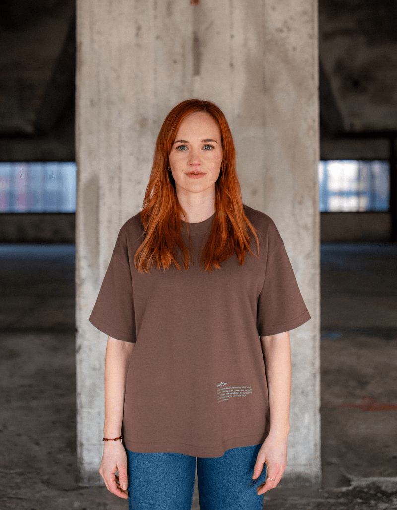 Frau mit roten Haaren trägt braunes T-Shirt mit kleinem Schriftzug unten rechts und steht vor einer Betonwand