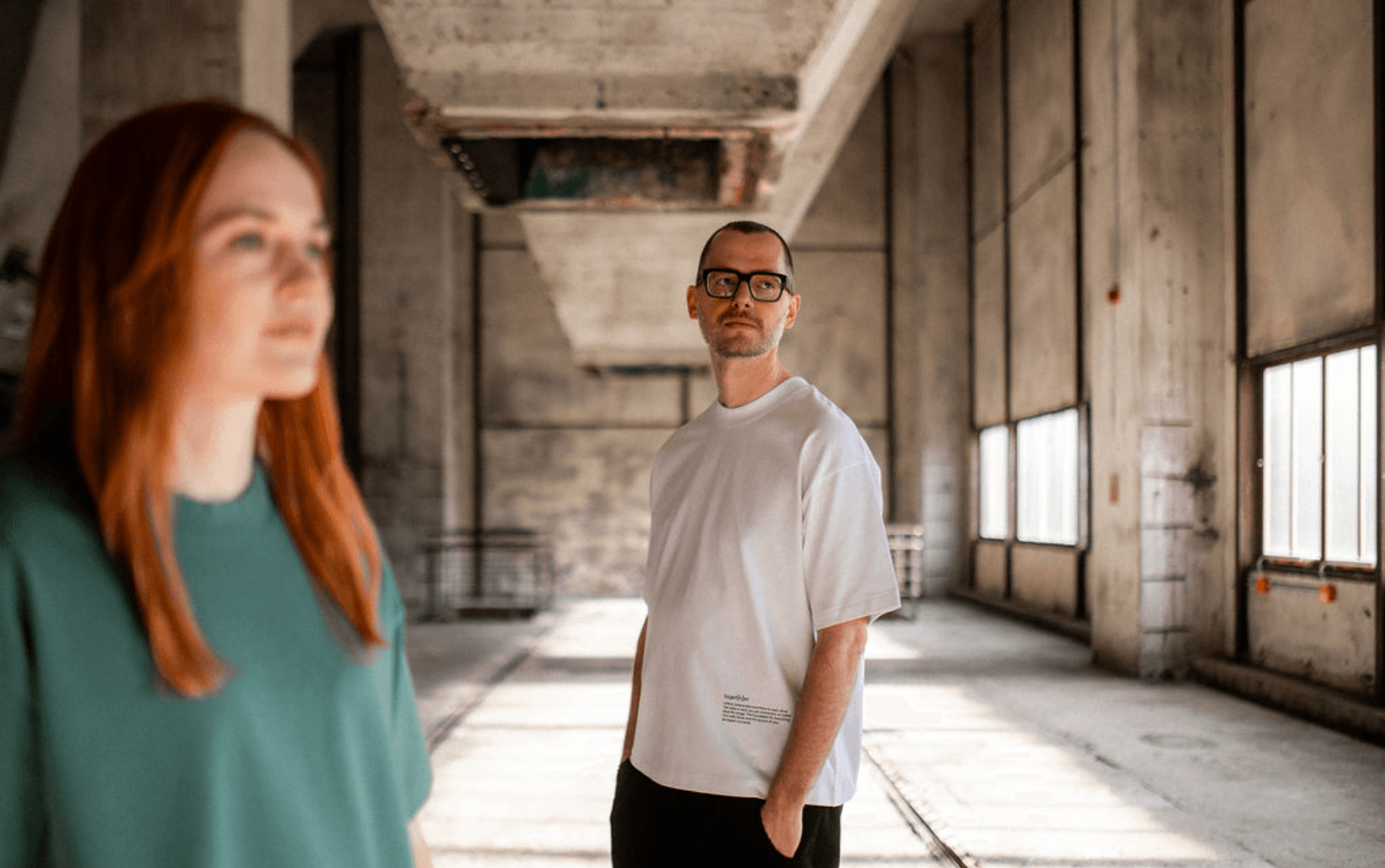 Person trägt weißes T-Shirt mit kleinem Schriftzug unten rechts und steht in einer hellen Industriehalle; im Vordergrund unscharf eine weitere Person