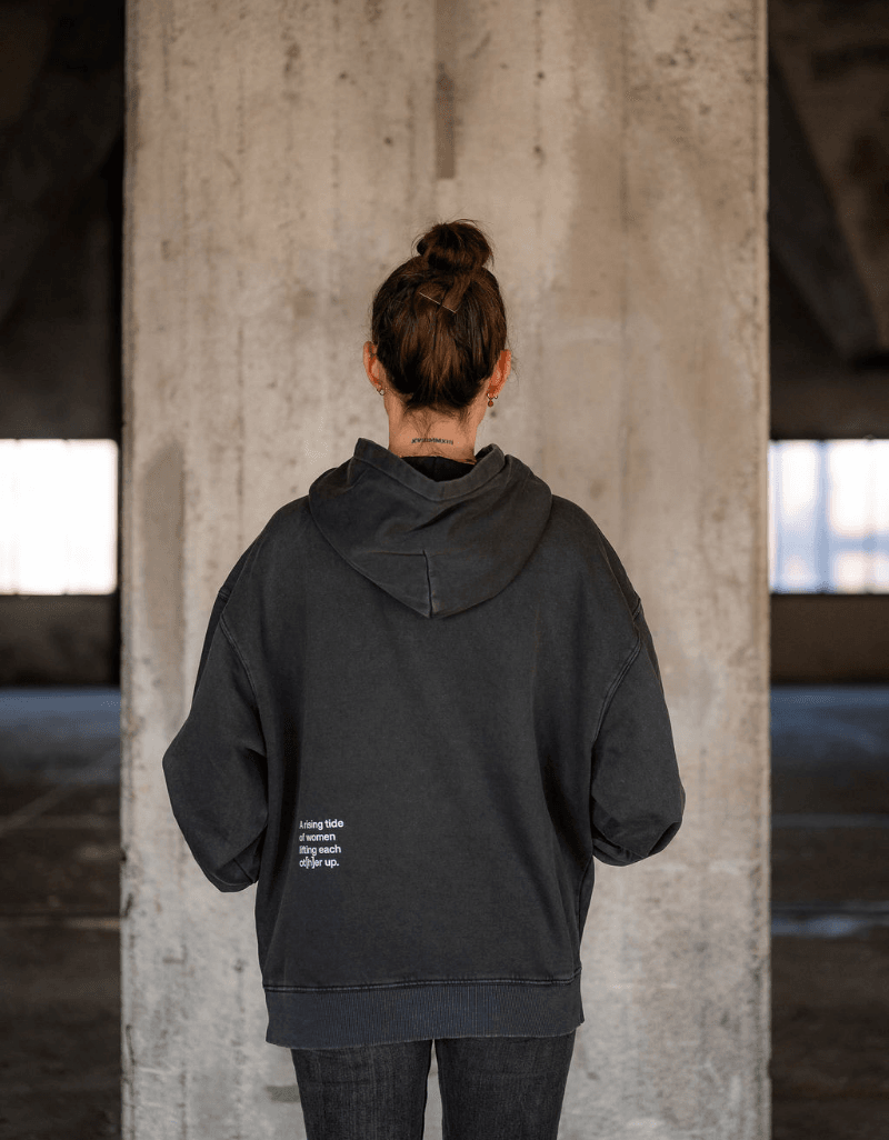 Person von hinten trägt schwarzen Hoodie mit kleinem Schriftzug im unteren Rückenbereich vor Betonwand