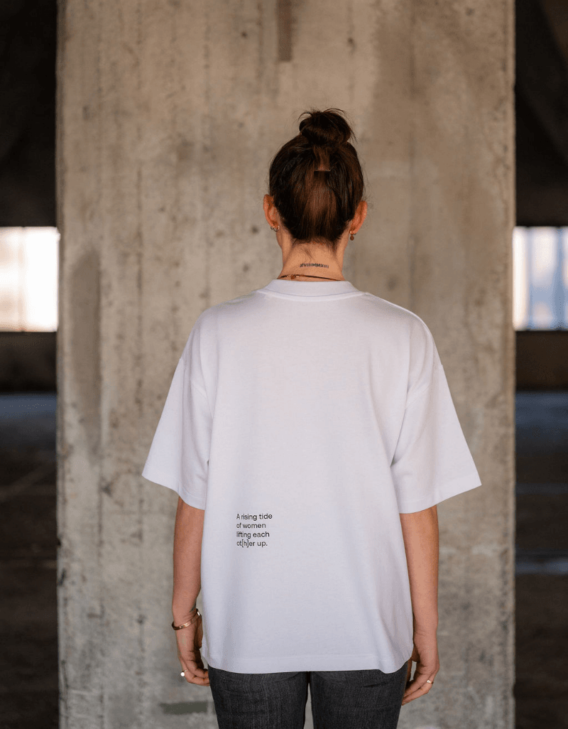 Person von hinten trägt weißes T-Shirt mit kleinem Schriftzug im unteren Rückenbereich vor Betonwand